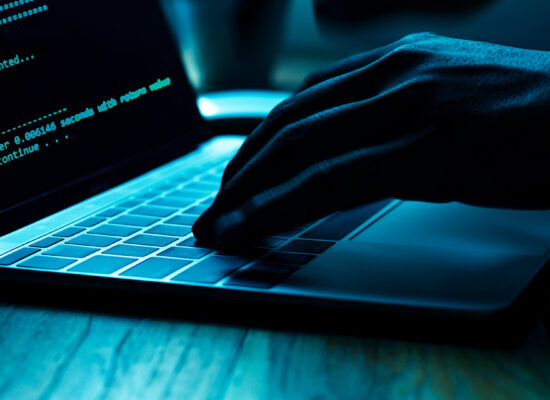 A close-up view of a person's hand typing on a laptop keyboard in a dimly lit setting. The screen displays green text resembling code or programming, creating a tech-focused atmosphere. The lighting emphasizes the hand and keyboard, suggesting cybersecurity or coding activity.